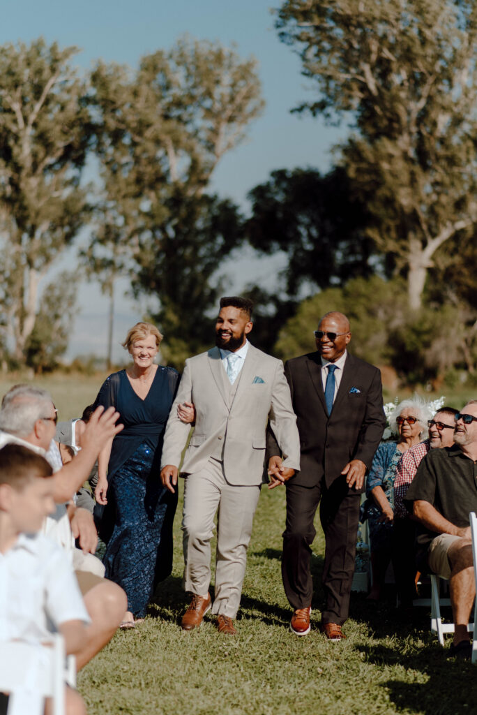 Groom walking down the outdoor ceremony aisle between two family members as guests look on.