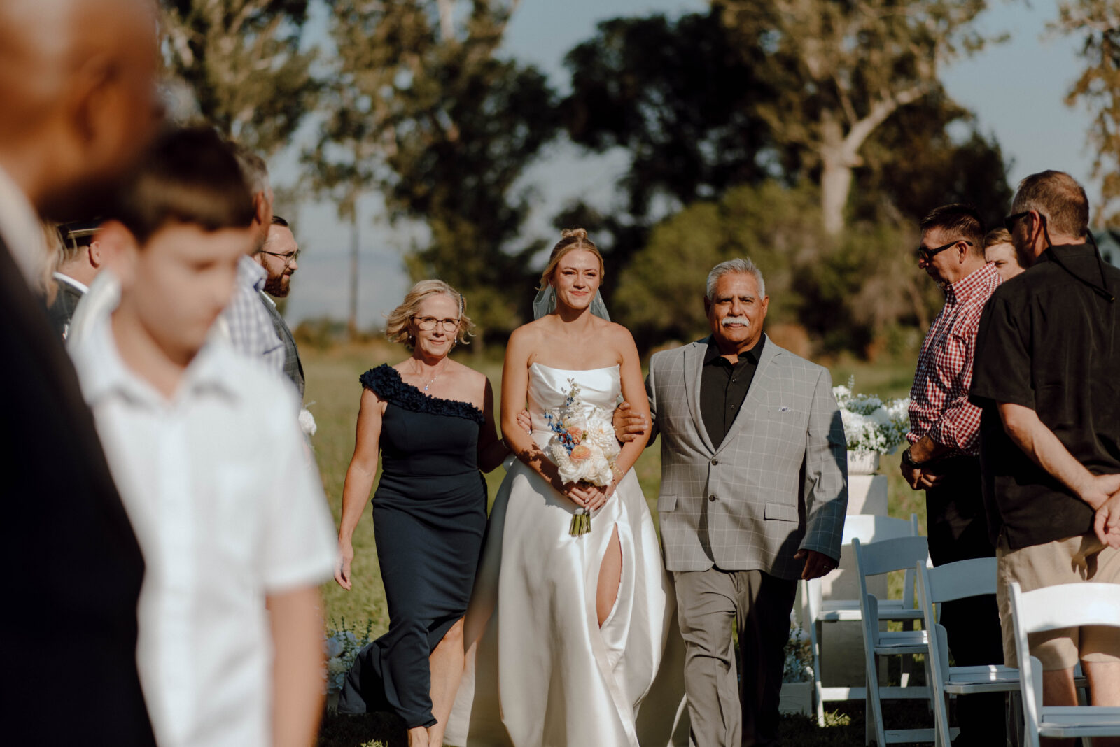 Bride walking down the grassy ceremony aisle between two family members.