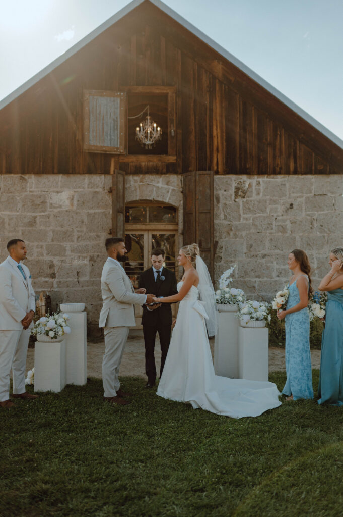 Bride and groom exchanging rings in front of the Stone Barn at Gansberg Ranch.