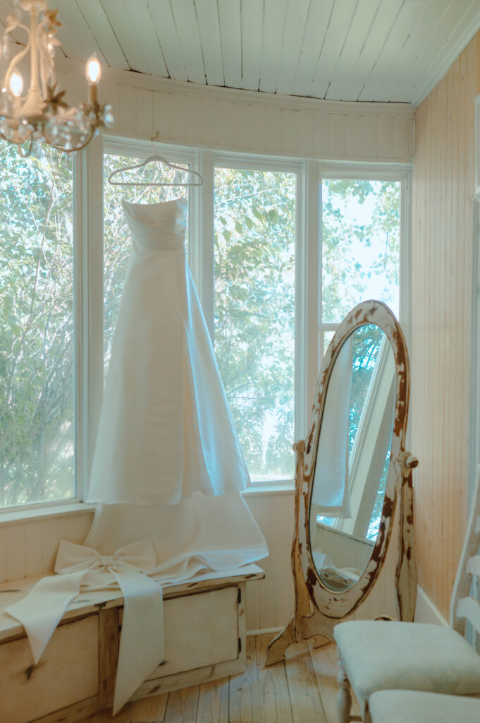 Wedding dress hanging in a bright bridal getting ready room beside a vintage mirror during a Gansberg Rnach wedding.