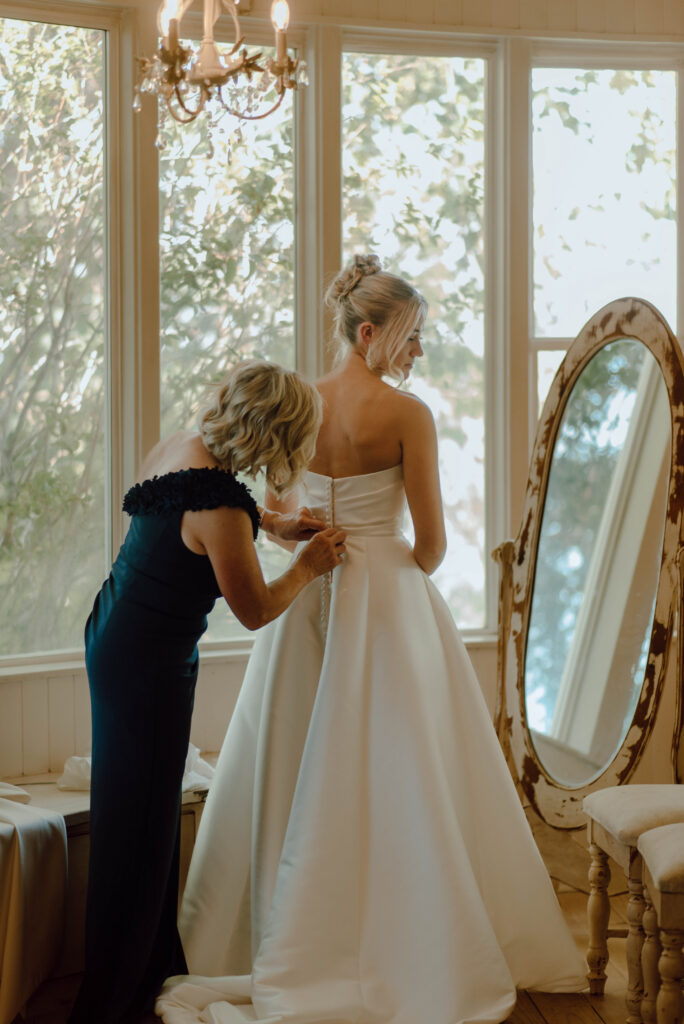 Bride getting into her wedding dress beside large windows and a vintage floor mirror during a Gansberg Rnach wedding.