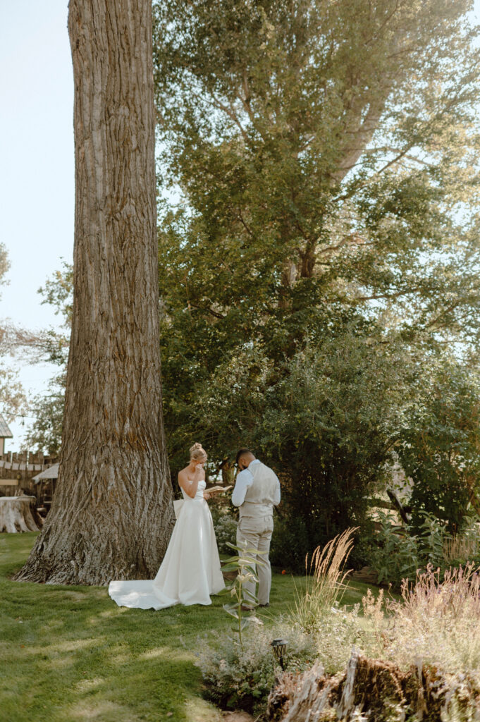 Bride and groom reading private vows together outside beneath a large tree.