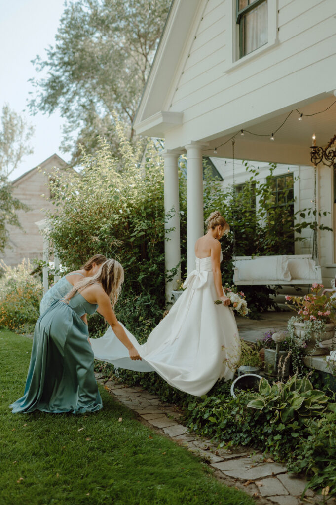 Bridesmaid holding the bride’s train as she walks toward the Gansberg Ranch house.
