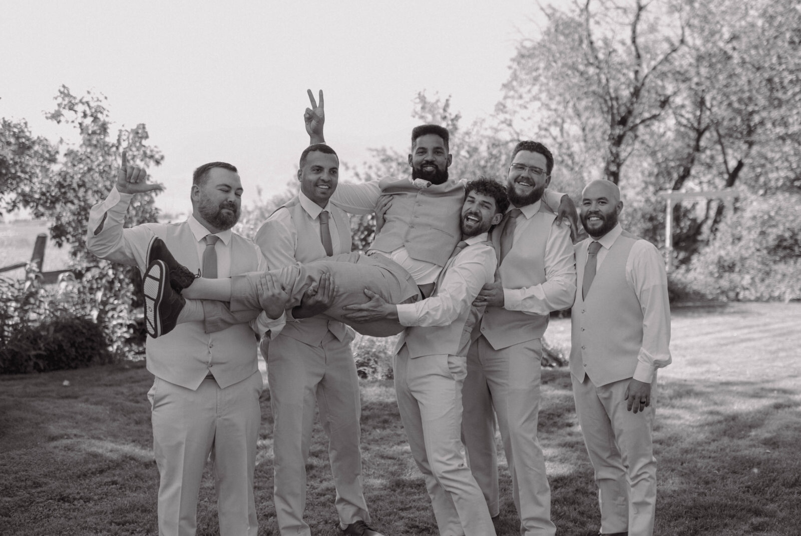 Black and white photo of groomsmen laughing while holding the groom horizontally.