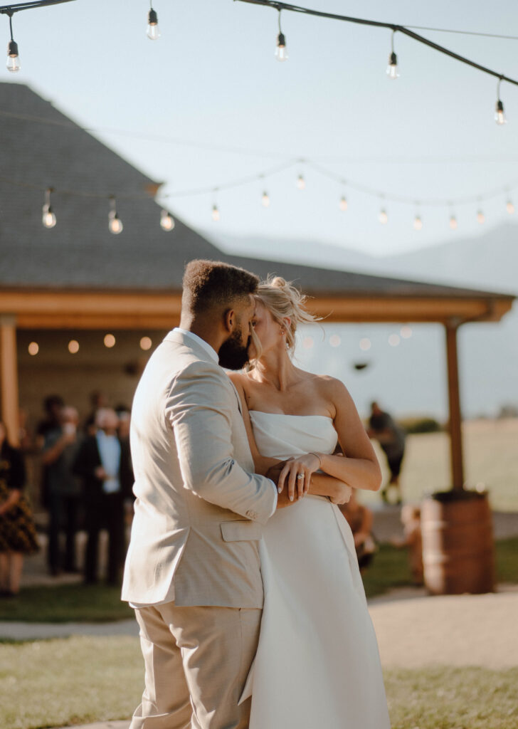 Bride and groom kissing during their first dance outdoors beneath string lights.