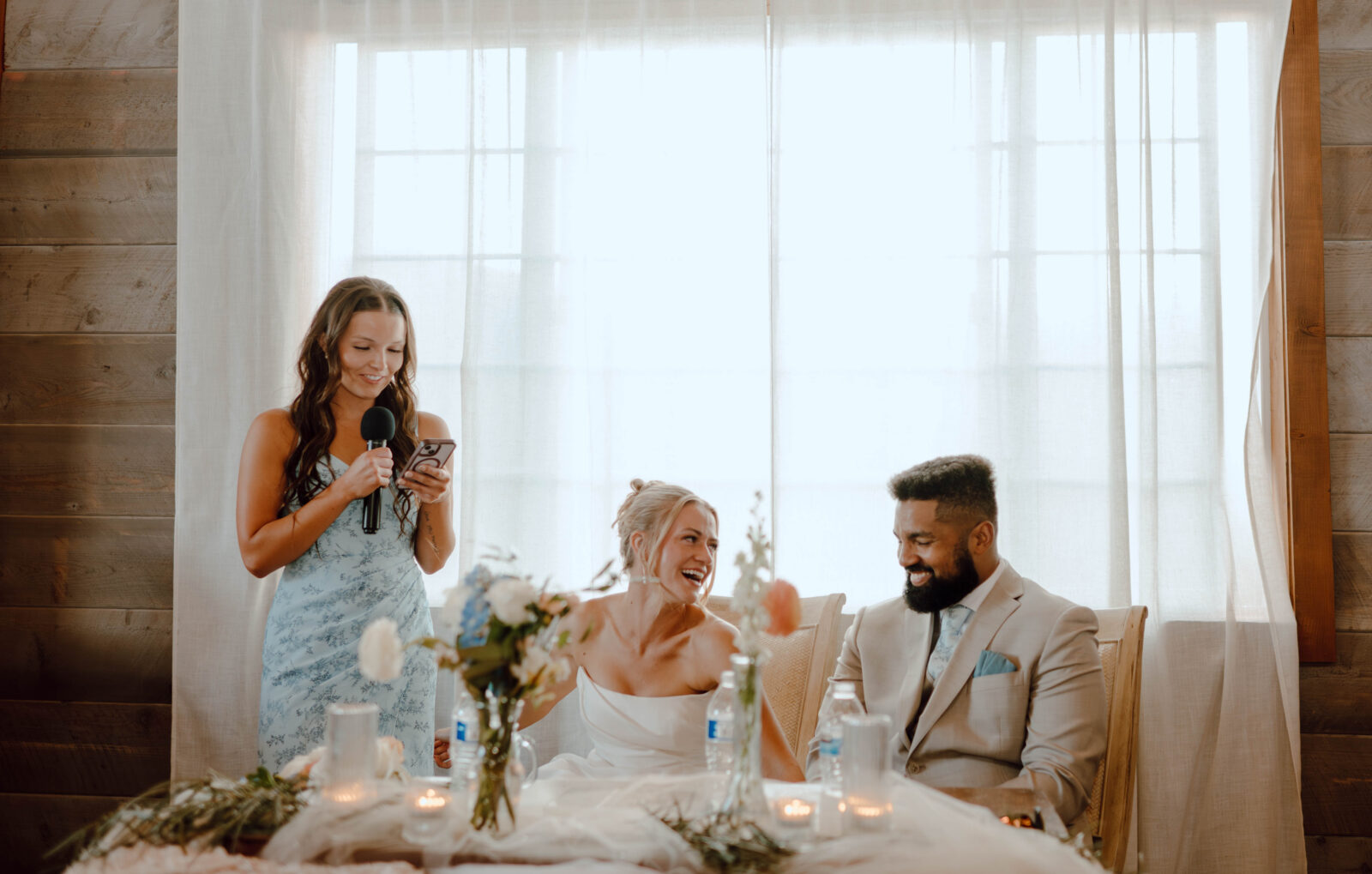 Bridesmaid giving a speech while the bride and groom laugh at their reception table.
