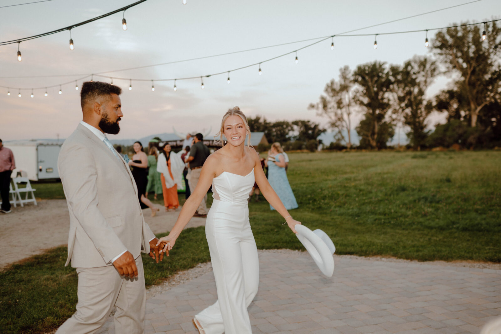 Bride in a white jumpsuit holding hands with the groom outside beneath string lights.