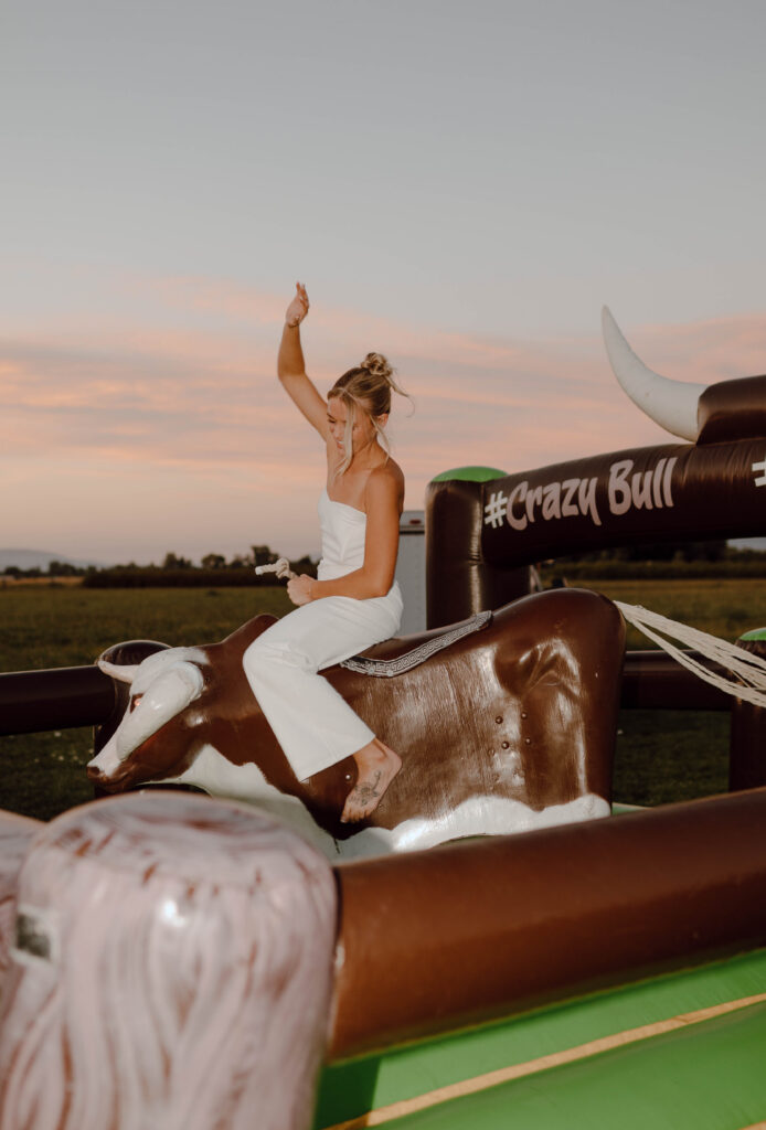 Bride riding a mechanical bull in her white reception jumpsuit at sunset.