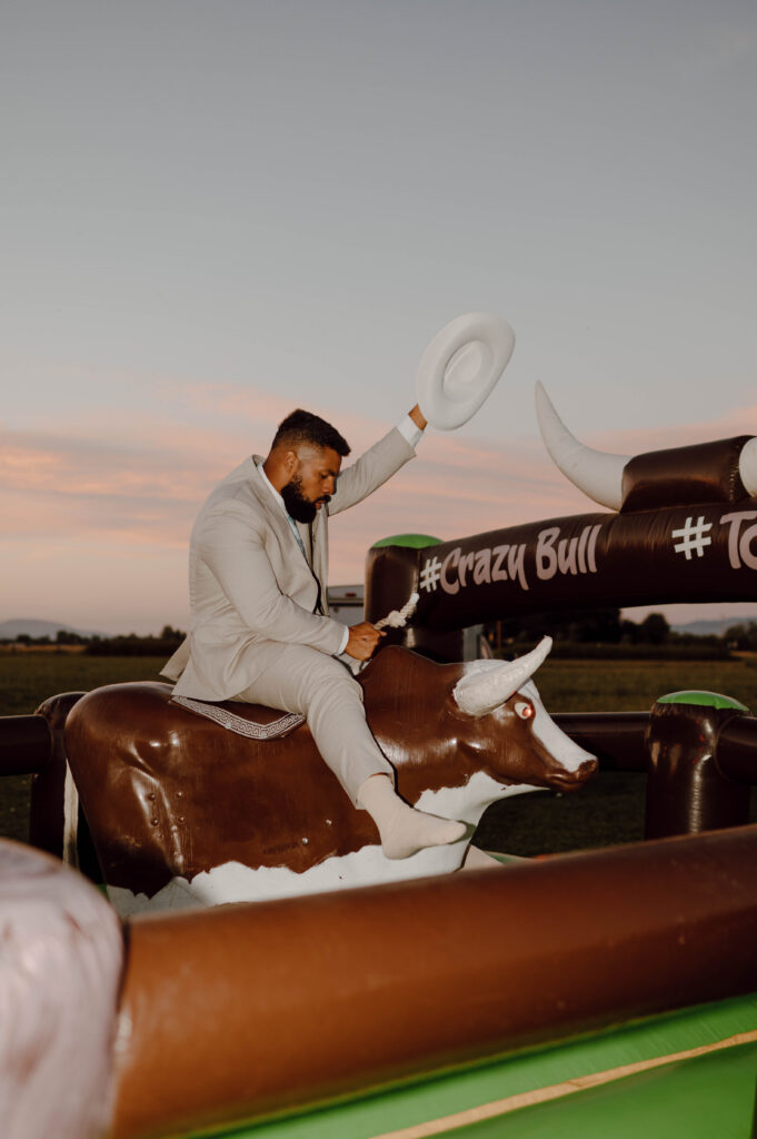 Groom riding a mechanical bull while holding a white cowboy hat in the air.