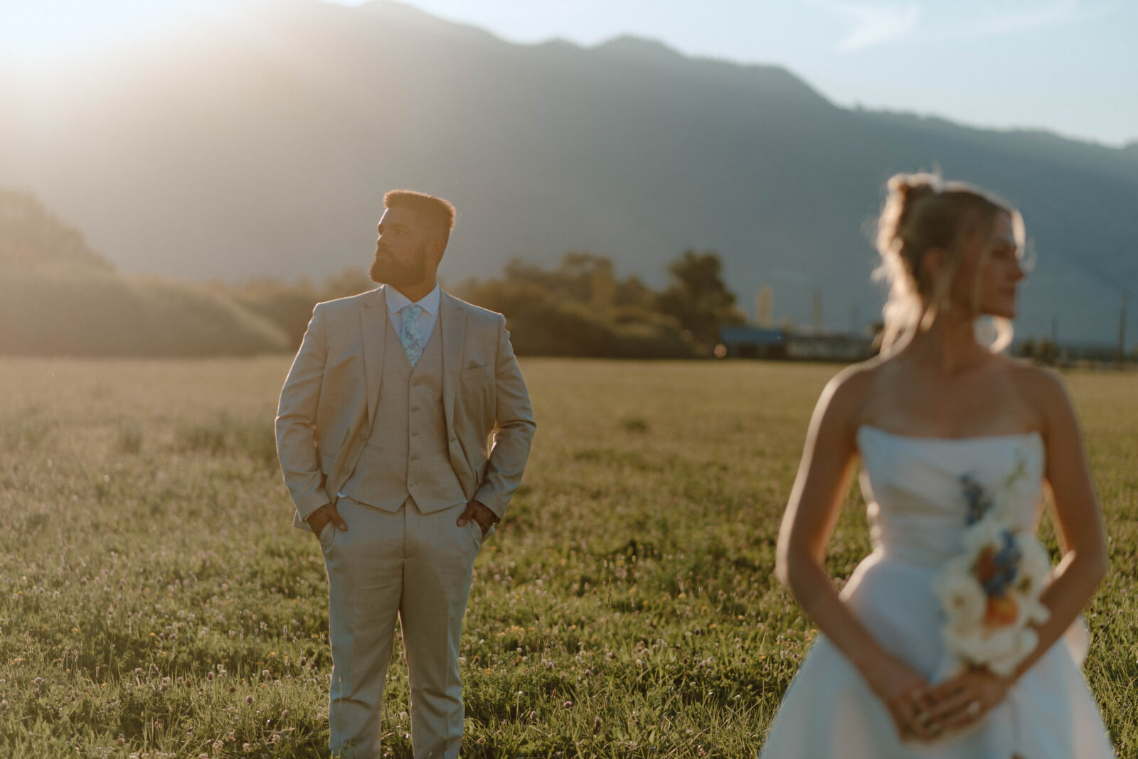 Bride and groom posing apart in a grassy field with mountains and golden evening light behind them.
