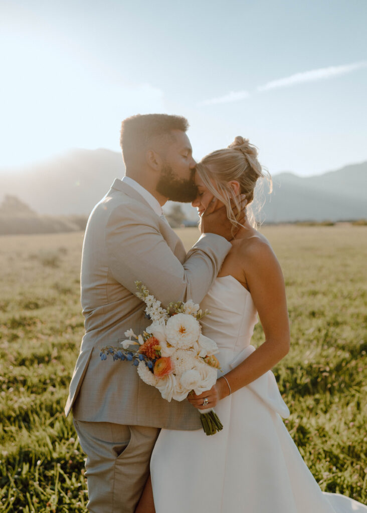 Groom kissing the bride’s forehead while she holds a white and blue wedding bouquet at golden hour.