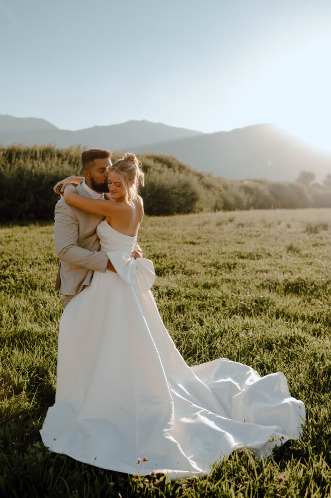 Bride and groom embracing in a sunlit field with mountains in the background.