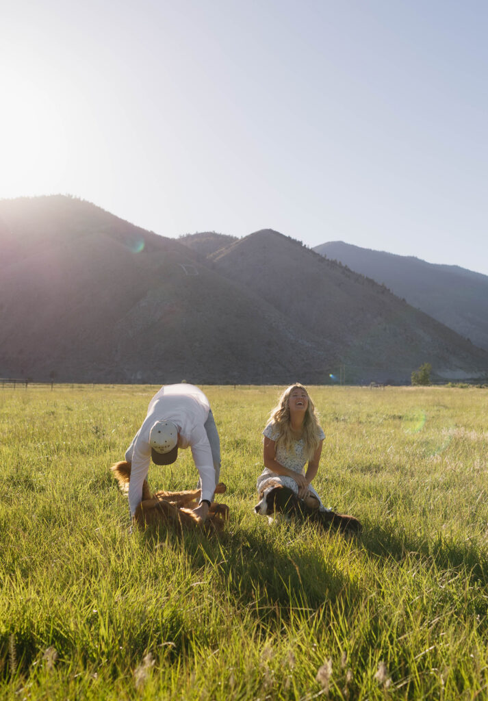 Candid engagement photo of a couple laughing as they play with their dogs in an open grassy mountain field. 