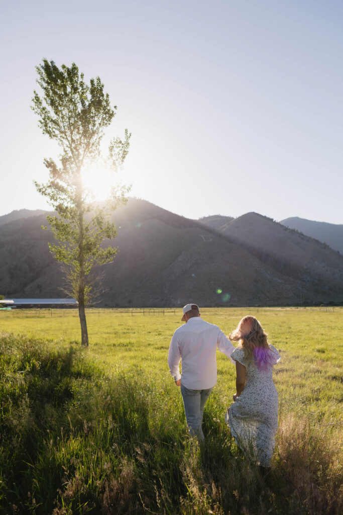 Couple walking towards a tree in an open field with the mountains in the background.