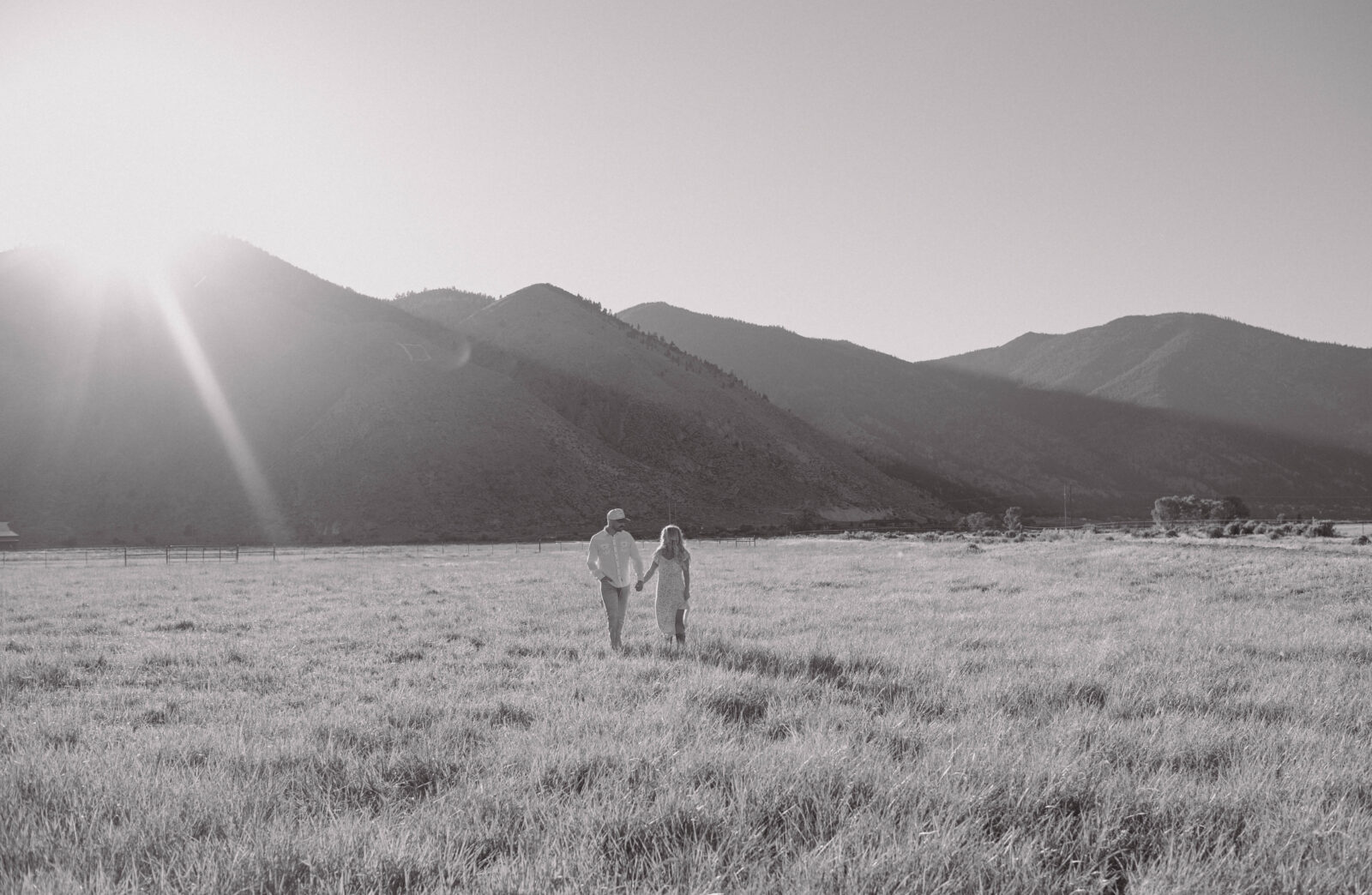 Black and white photo of a couple walking in a grassy field with mountains behind them.