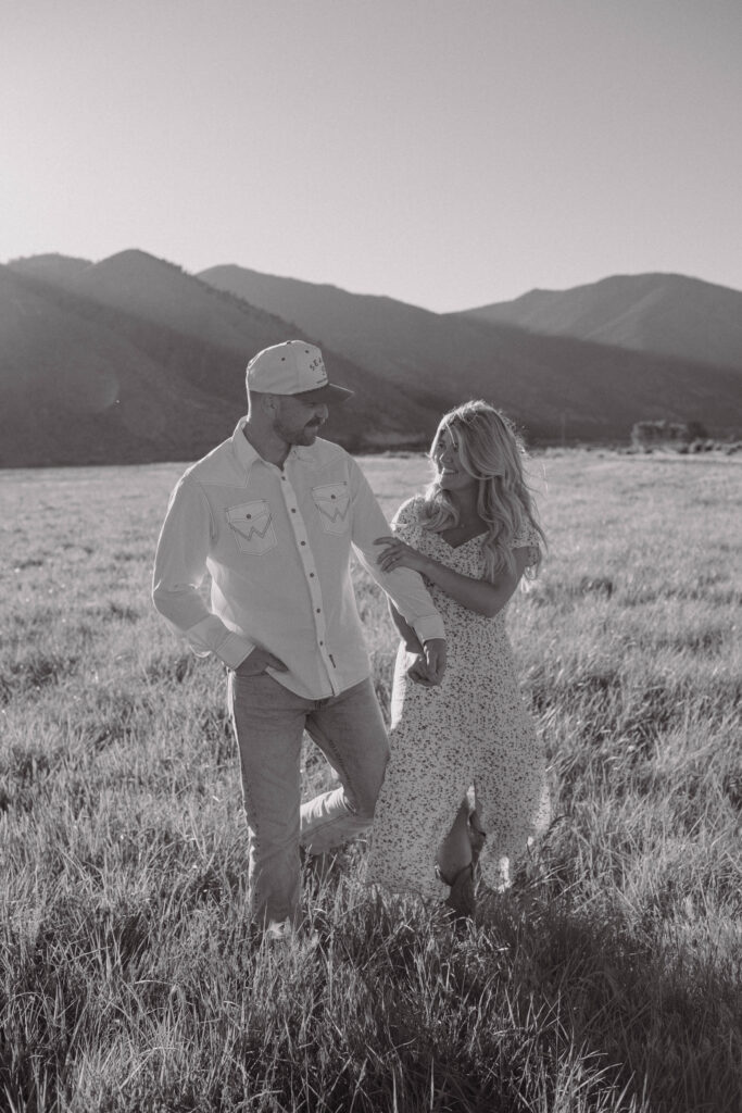Black and white photo of a couple walking in a grassy field with mountains behind them.