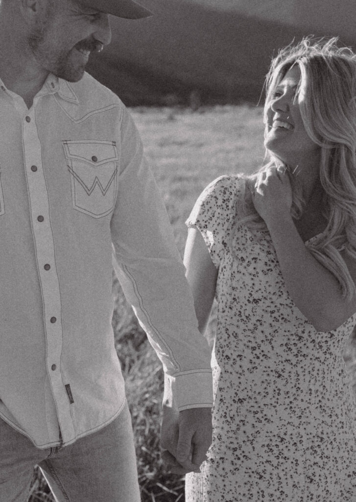 Black and white photo of a couple smiling at one another and holding hands in a grassy field. 