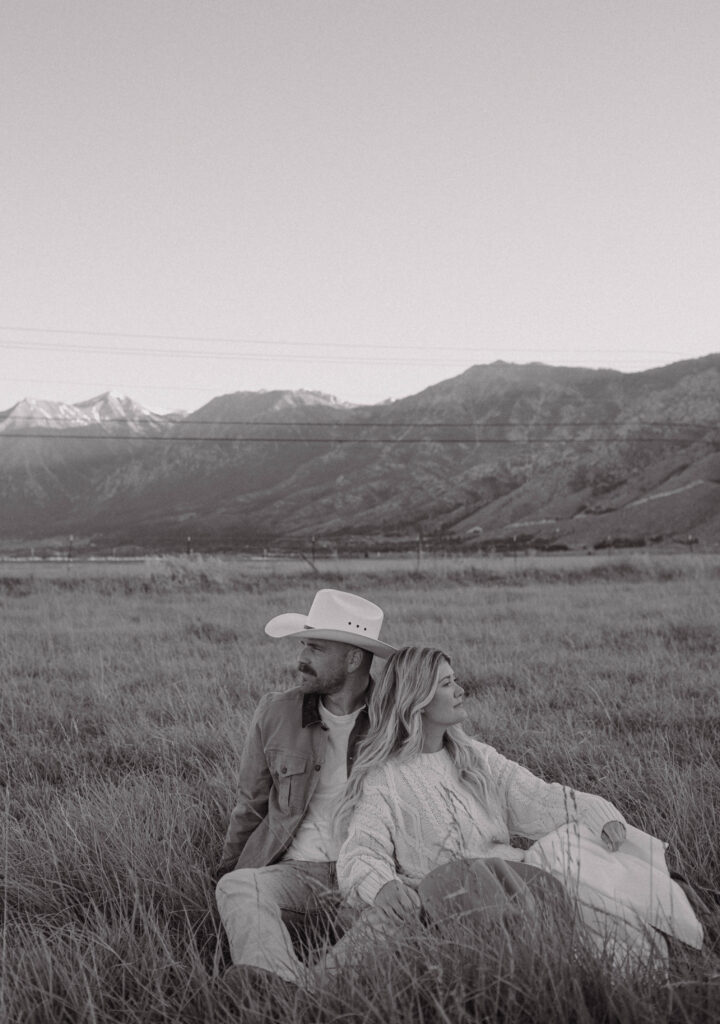 Black and white photo of a couple sitting in the grass during their mountain engagement photos in Nevada with Jobs Peak in the background. 